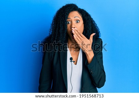 Middle age african american woman using lavalier microphone covering mouth with hand, shocked and afraid for mistake. surprised expression 