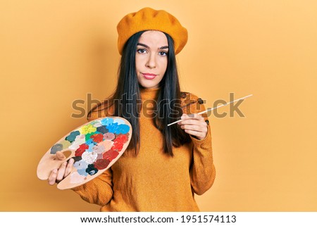 Young brunette woman holding paintbrush and palette wearing beret relaxed with serious expression on face. simple and natural looking at the camera. 