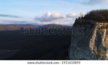 Golden cross on a top of a mountain with steep slope, concept of religion. Shot. Aerial view of a cliff and beautiful hilly valley on the background.