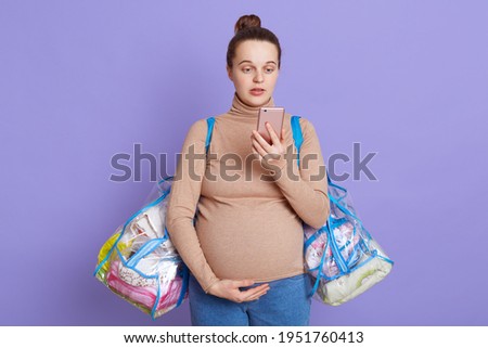 Pregnant beautiful woman posing with two bags and holding smart phone in hands, looking at display with puzzled loo, need to call and go to maternity home, posing isolated over lilac background.