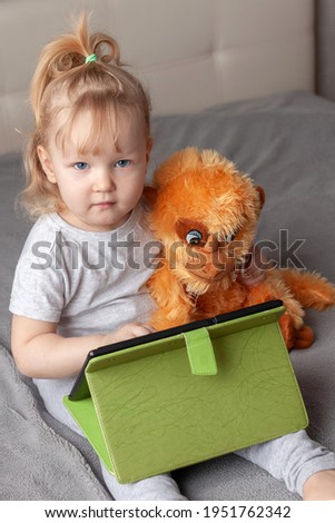 preschool girl plays tablet home with soft toy, educational concept for school children. blue-eyed blonde looks camera