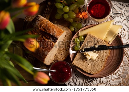 
Breakfast is ready! Bread is sliced, smeared with butter, glass of tea, grapes, tulips and strawberry jam stand next to it on the wooden table decorated with a white tablecloth. Rustic