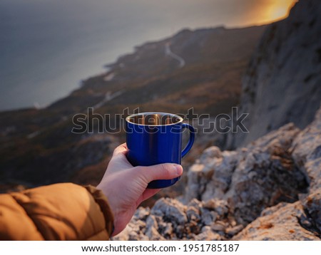 Closeup photo of cup with tea in traveler's hand over out of focus mountains view. A tourist man drink a hot tea from cup and enjoys the scenery in the mountains. Trekking concept