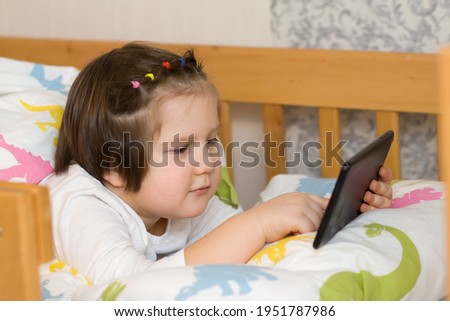 Cute Girl  lying on a soft bed. Child watching cartoons on touchpad. Playing games Kids are studying online with black tablet. Children use technology for education and satiations