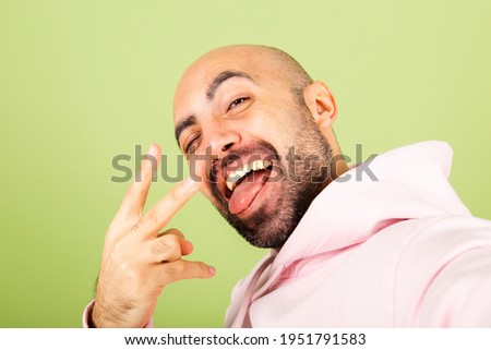 Young bald caucasian man in pink hoodie isolated on green background positive cheerful take photo selfie