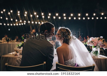 A young groom in a gray suit and a smiling blonde bride sit with their backs at the table on chairs, look at each other and laugh, against the background of electric bulbs, garlands. Night portrait. Royalty-Free Stock Photo #1951832164