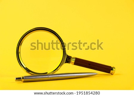 A magnifying glass in a gold frame and a pen on a yellow background. Copy space.