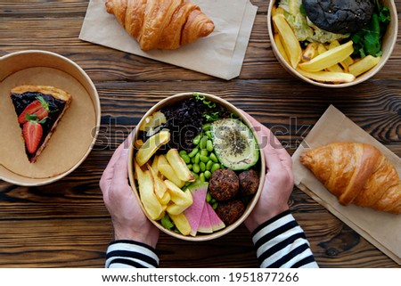 Pov shot of man holding hawaiian vegan poke bowl over the wooden table, takeout food. French croissant, salmon burger with black bun, blueberry cheesecake. Close up, top view, copy space, background