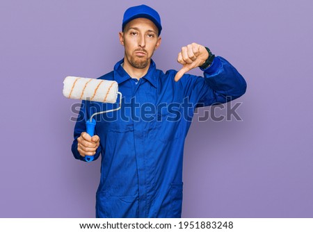 Bald man with beard holding roller painter with angry face, negative sign showing dislike with thumbs down, rejection concept 