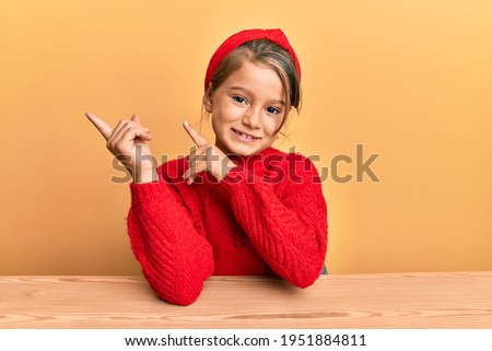Little beautiful girl wearing casual clothes sitting on the table smiling and looking at the camera pointing with two hands and fingers to the side. 