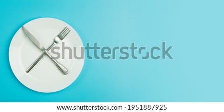 Empty plate on a blue minimal banner background. Empty white ceramic plate with knife and fork on the table after eating. Diet and healthy food concept. Royalty-Free Stock Photo #1951887925