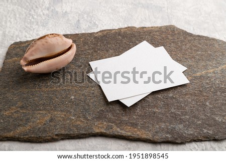 White paper business card, mockup with natural stone and seashell on gray concrete background. Blank, side view, still life, copy space.