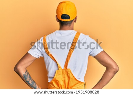 Young handsome african american man wearing handyman uniform over yellow background standing backwards looking away with arms on body 