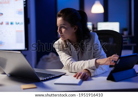 Manager woman using laptop and tablet in same time working on financial reports in business start-up office . Busy multitasking employee using modern technology network wireless late at night Royalty-Free Stock Photo #1951916410