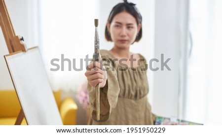 Creative influencer or female blogger an online streaming painting demonstration. A young woman Asian artist holding a paint palette and paint brushes is broadcasting live with a smartphone at home.