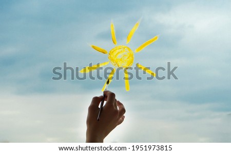 Children's hand draws with paints the sun on the glass, against the background of the cloudy sky