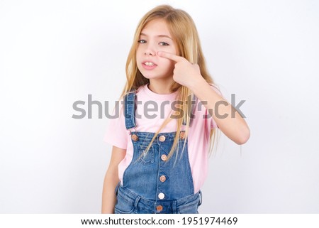 beautiful caucasian little girl wearing jeans overall over white background pointing unhappy to pimple on forehead, ugly infection of blackhead. Acne and skin problem