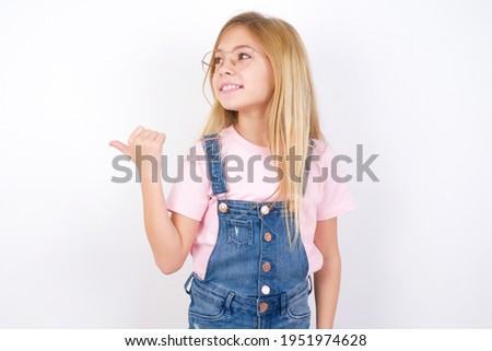 Charming beautiful caucasian little girl wearing jeans overall over white background looking at copy space having advertisements