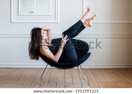 A brunette girl in black leggings lies on a black chair and does yoga exercises. Background white