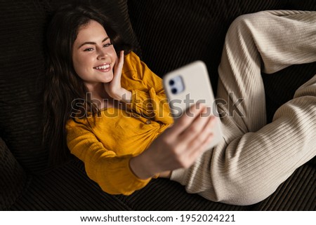 Smiling young brunette woman using mobile phone while laying on a couch, taking selfie