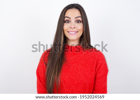 young beautiful brunette excited woman wearing red knitted sweater over white wall smiling to the camera after hearing amazing news from her family or friends 