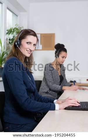A young business woman with headset is typing at a notebook. Another woman with headset is sitting in the background (blurred).