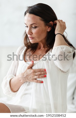 Middle aged woman sitting on a white bed in a bedroom at home touching chest with her hands while having a heart pain and feeling unwell. Suffering from stress, hypertension or cardiac arrhythmia. Royalty-Free Stock Photo #1952108452