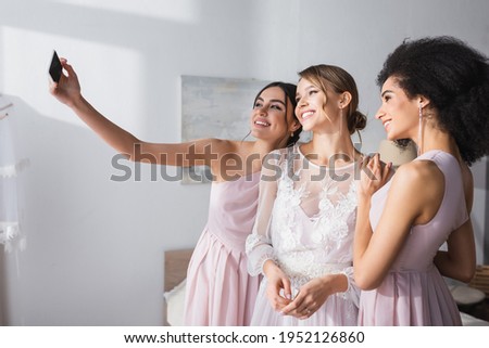 cheerful woman taking selfie with bride and african american friend in bedroom