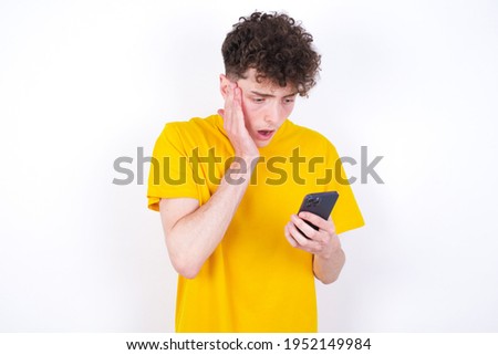young caucasian handsome man with curly hair wearing yellow T-shirt against white studio background holding in hands cell reading sms using new app 5g
