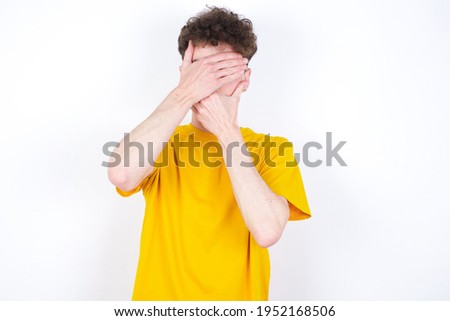 young caucasian handsome man with curly hair wearing yellow T-shirt against white studio background Covering eyes and mouth with hands, surprised and shocked. Hiding emotions.