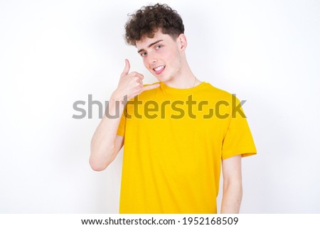 young caucasian handsome man with curly hair wearing yellow T-shirt against white studio background smiling doing phone gesture with hand and fingers like talking on the telephone. Communicating