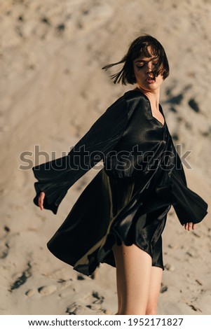 A young attractive dark-haired model girl posing and dancing in the pine forest in front of the sand quarry. Female model wearing black dress outdoors.