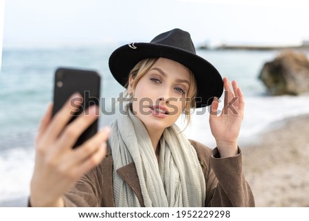 Portrait of stunning female model taking a selfie using her mobile phone while holding a stylish hat 