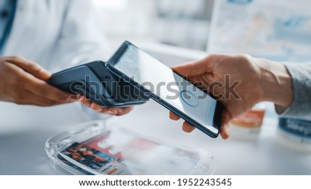 Pharmacy Drugstore Checkout Cashier Counter: Pharmacist and a Customer Using NFC Smartphone with Contactless Payment Terminal to Buy Prescription Medicine, Health Care Goods. Close-up Shot Royalty-Free Stock Photo #1952243545
