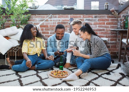 Stylish mixed race friends relaxing together on rooftop with alcoholic cocktails and tasty pizza. Happy young people sitting on floor and using modern smartphone.