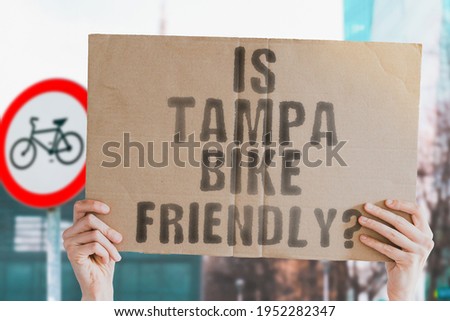 The question " Is Tampa bike friendly? " on a banner in men's hand with blurred background. Transportation. Zero waste. Bicycle lane. Streets. City. Safety. Insecure. Road signs. Dangerous