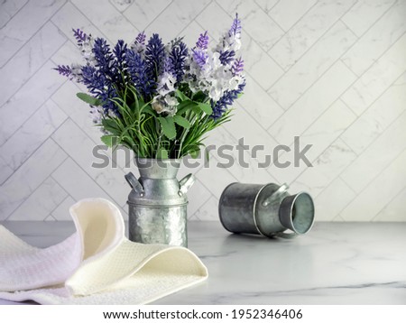 Rustic metal milk can filled with artificial purple lavender and lilacs for a simple spring country home decor with still life photography and a marble counter with a herringbone tile background.  