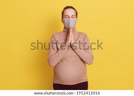 Attractive young European woman posing with closed eyes, keeping palms together in front of chest, praying heartily to god, pleading forgiveness, isolated over yellow background.
