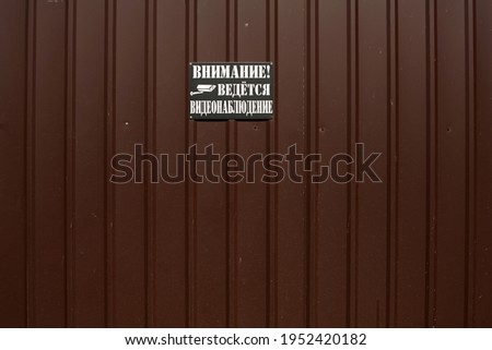 A plate hangs on a metal fence with a brown profile. The caption reads "Attention, video surveillance is underway" 