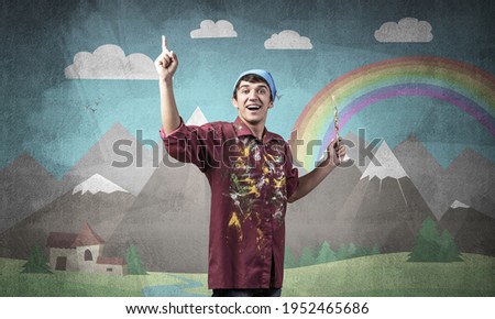 Young male artist with paintbrush. Happy painter in shirt and bandana standing on background colorful picture on wall. Summer landscape with mountains and rainbow. Creative hobby and profession