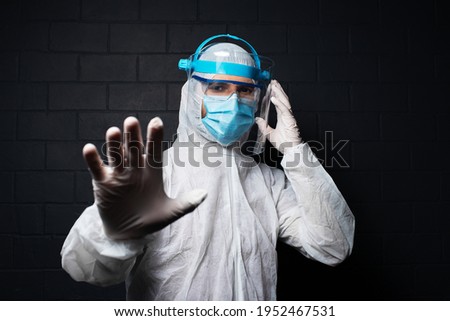 Dark studio portrait of young doctor wearing PPE suit against coronavirus and covid-19. Showing stop sign with hand. Background of black brick wall.