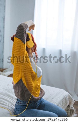 A girl with red bright hair sits on the bed, looks out the window and listens to music in yellow headphones.
