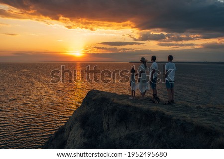 mom dad daughter and adult son in white clothes stand on a hill by the sea at sunset rear view