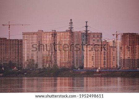Construction of multi-storey buildings and cranes on the embankment of the river, against the background of a purple evening sky.