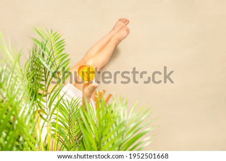 Alone woman sitting under palm tree branches with glass of water with piece orange. Female relaxation on the sand of the beach at summer vacation. Top view
