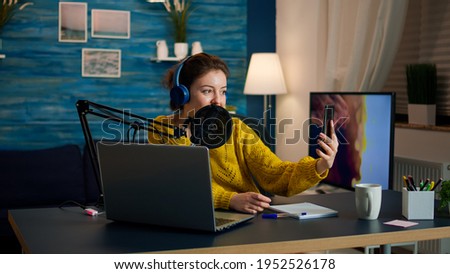 Influencer with headphones using phone for taking selfie recording podcast series for audience. On-air online production internet broadcast show host streaming live content for digital social media
