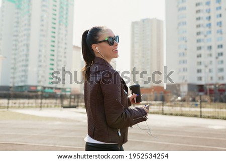 Wonderful pretty lady wearing sunglasses and leather jacket drinking coffee and listening music while walking in the city 