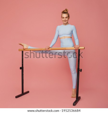 Young motivated determined woman in sportswear  leggings stretching legs on ballet barre standing on pink background focused exercising, preparing muscles working-out
