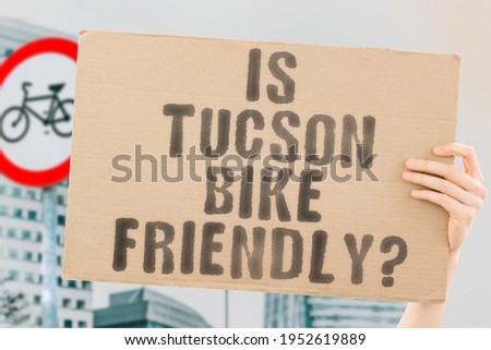The question " Is Tucson bike friendly? " on a banner in men's hand with blurred background. Transportation. Zero waste. Bicycle lane. Streets. City. Safety. Insecure. Road signs. Dangerous