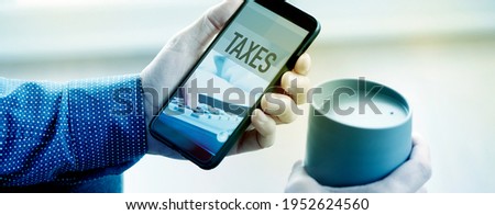 a young man, wearing a blue shirt, has his smartphone in one hand, with the text taxes in its screen, and a cup of coffee in the other hand, in a panoramic format to use as web banner or header